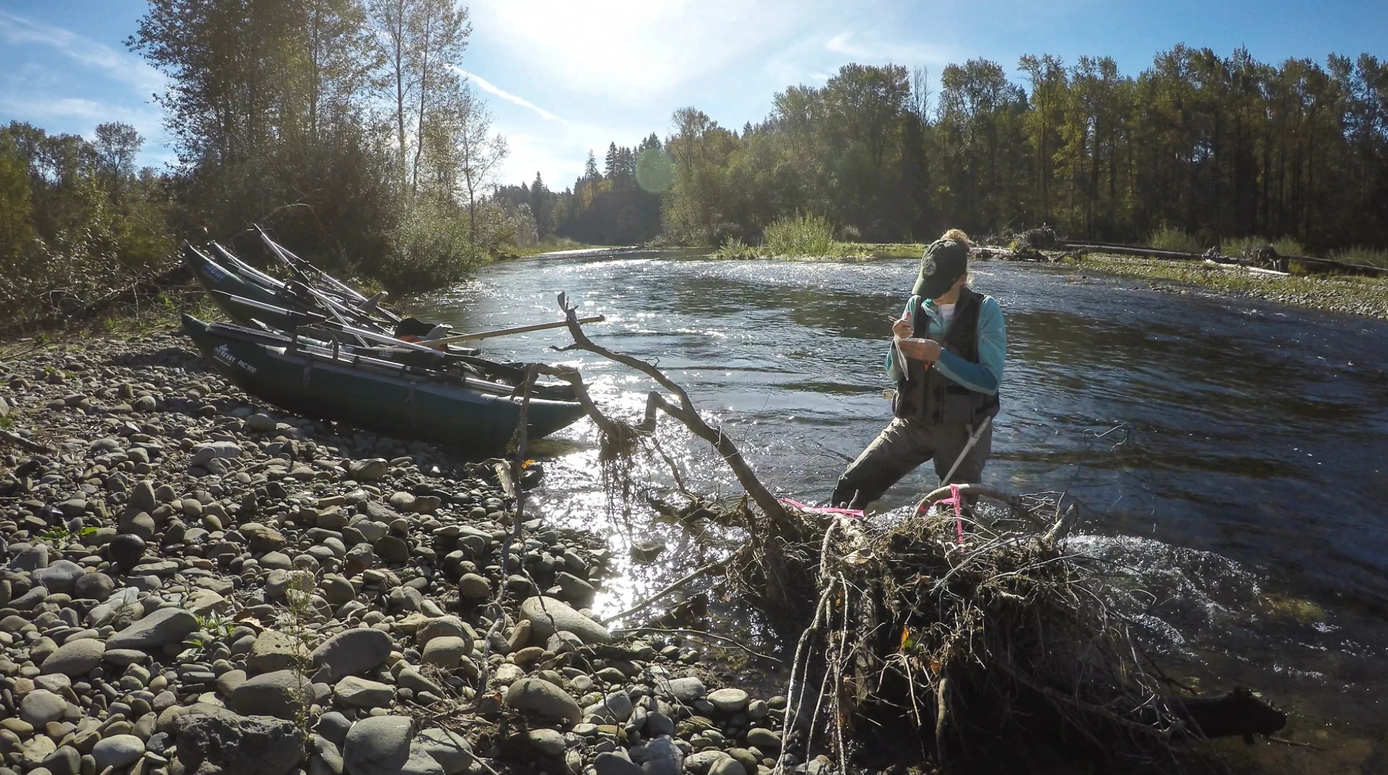 A field technician wearing waders stands in a shallow, rocky river near the bank, taking notes while stabilizing themselves on a partially submerged log. A small survey boat loaded with gear rests on the gravel shoreline behind them, with trees lining the river under a sunny sky.